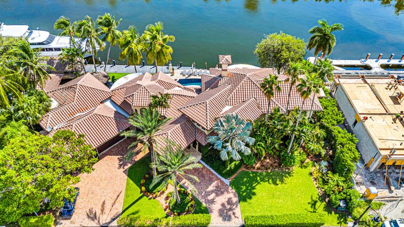 Sunlit aerial view of a luxury waterfront Mediterranean-style mansion with terracotta tile roof, palm-lined driveway, manicured lawn, tropical landscaping and private dock with a yacht along a calm canal