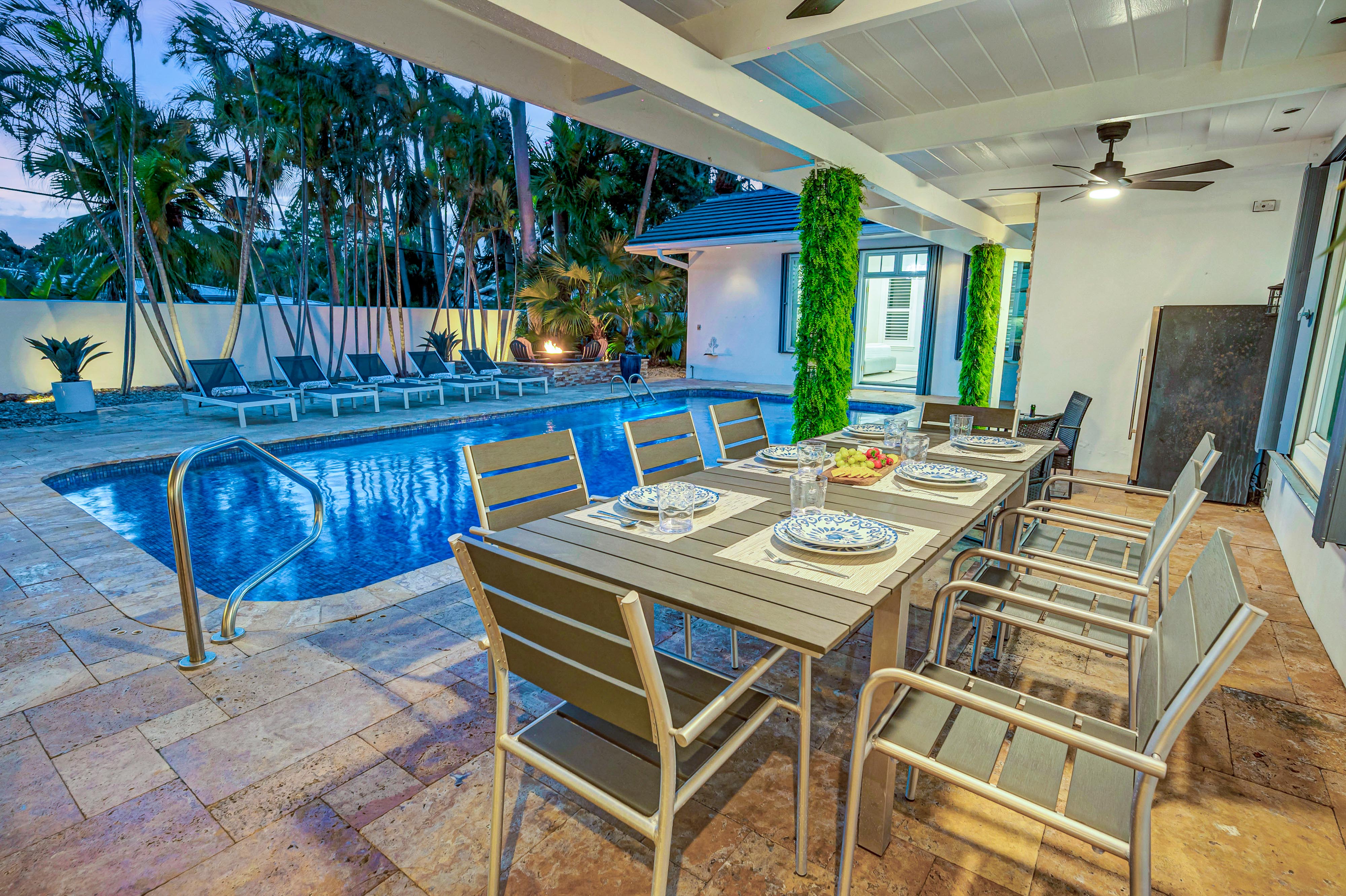 Dusk-lit tropical backyard patio with covered outdoor dining table set for eight, ceiling fan, stone tile deck and illuminated swimming pool with lounge chairs.