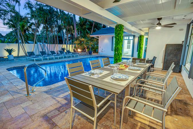 Dusk-lit tropical backyard patio with covered outdoor dining table set for eight, ceiling fan, stone tile deck and illuminated swimming pool with lounge chairs.