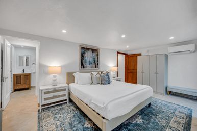 Bright coastal king bedroom with crisp white linens, blue patterned area rug, matching nightstands and lamps, wardrobe and bench, wall-mounted AC, and adjacent en-suite bathroom.