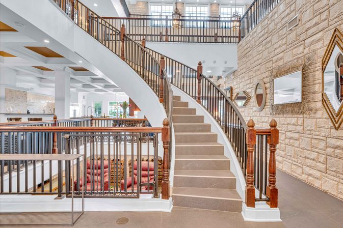 Bright modern interior lobby with a sweeping curved staircase and wooden banister, stone accent wall with decorative mirrors, mezzanine railing overlooking red lounge seating