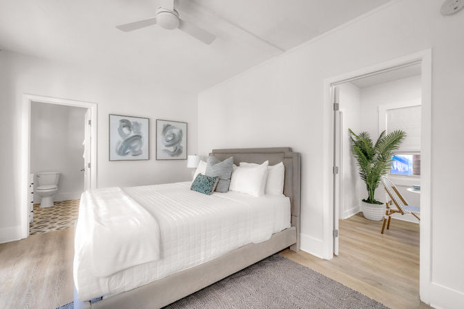 Bright modern bedroom with gray upholstered bed and crisp white linens, abstract blue wall art, ceiling fan, doorway to an en-suite bathroom with patterned tile, and an adjacent sunlit reading nook with a potted palm and wicker chair.