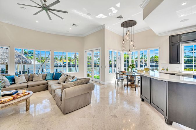 Sunny open-concept waterfront living room and kitchen with large windows overlooking a tropical canal, beige sectional sofa, marble floors, island bar and modern pendant lights.