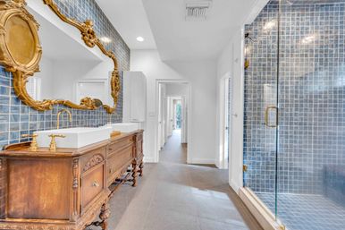 Bright luxury master bathroom with blue mosaic tile wall, glass walk-in shower, antique carved wooden double vanity with white vessel sinks and gold faucets, and ornate gilt mirror.