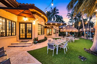Cozy sunset-lit tropical backyard patio at a Mediterranean-style villa with string lights, palm trees, thatched umbrellas and tables set for al fresco dining on a manicured lawn.