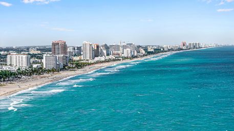 Aerial view of a sunny beachfront city with high-rise hotels and condos lining a sandy shore, turquoise Atlantic waves and blue sky.