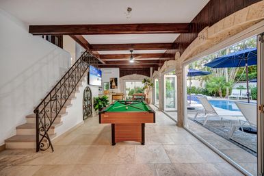Indoor-outdoor poolside game room in a luxury home: green pool table under dark wood beams, wrought-iron staircase, stone archways and tile floor opening via sliding glass doors to a tropical pool patio with lounge chairs and blue umbrellas.