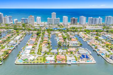 Aerial view of a tropical waterfront canal neighborhood with luxury homes, private docks and pools, palm-lined streets and oceanfront high-rise condos along the turquoise Atlantic coast.