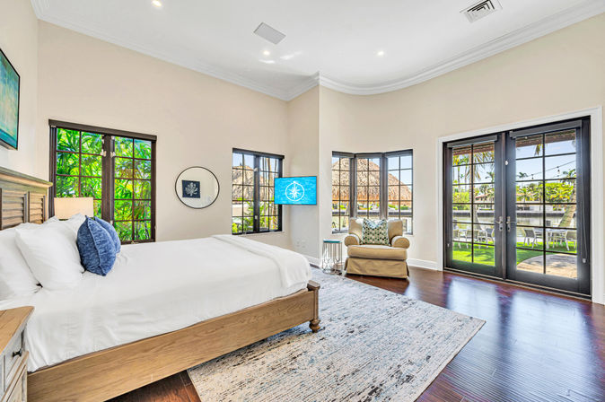 Sunlit tropical waterfront bedroom with king bed, hardwood floors, bay windows and French doors opening to a grassy patio and canal, cozy armchair, white linens and blue accent pillows in modern coastal decor.