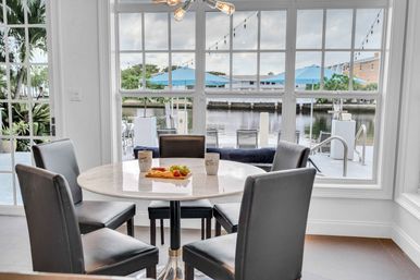 Bright waterfront breakfast nook with round marble table and four black chairs, coffee mugs and fruit platter on the table, large windows framing a marina/canal view with blue umbrellas and dock