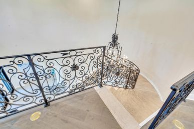 Top-down view of a curved interior staircase in a luxury home featuring ornate wrought-iron scrollwork railing, hanging chandelier, and light wood steps.