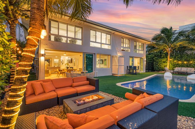 Sunset-lit tropical backyard of a luxury modern two-story home with an illuminated pool, orange cushioned patio sofa around a fire pit, palm trees wrapped in string lights, and open-plan indoor living visible.