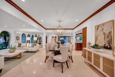 Bright open-concept dining and living area with marble floors, modern gold chandelier, arched windows revealing palm trees in a tropical villa-style interior.