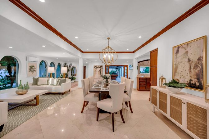 Bright open-concept dining and living area with marble floors, modern gold chandelier, arched windows revealing palm trees in a tropical villa-style interior.