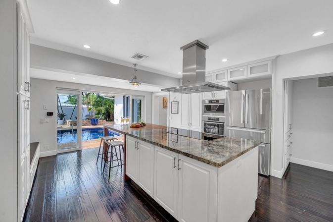 Modern open-concept kitchen with large granite island and breakfast bar, white cabinetry, stainless steel appliances and dark hardwood floors, opening through sliding glass doors to a sunny backyard pool and tropical plants.
