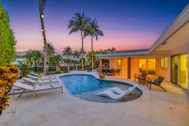 Inviting tropical waterfront backyard with curved swimming pool, in-pool loungers, sunbeds, palm trees, string lights and covered patio dining area at sunset