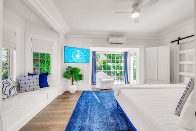 Sunny coastal-style bedroom with white walls and ceiling fan, king bed with blue accents, built-in window seat, blue rug, TV and sliding doors to a lush tropical garden patio.