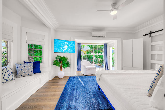 Sunny coastal-style bedroom with white walls and ceiling fan, king bed with blue accents, built-in window seat, blue rug, TV and sliding doors to a lush tropical garden patio.