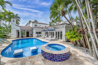 Luxury tropical backyard with blue swimming pool and raised mosaic spa, palm trees, sun loungers with umbrellas, and white stucco villa