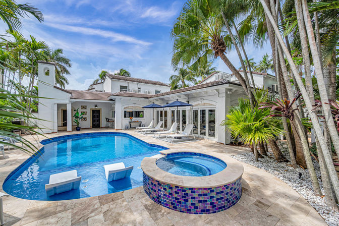 Luxury tropical backyard with blue swimming pool and raised mosaic spa, palm trees, sun loungers with umbrellas, and white stucco villa