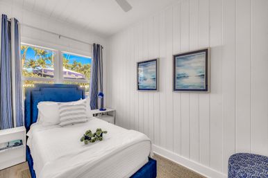 Sunny coastal bedroom with white shiplap walls, navy-blue upholstered bed and striped curtains, crisp white linens with decorative pillow, ocean-themed framed prints, blue bedside lamp, and palm trees visible through the window