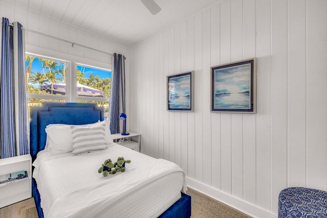 Sunny coastal bedroom with white shiplap walls, navy-blue upholstered bed and striped curtains, crisp white linens with decorative pillow, ocean-themed framed prints, blue bedside lamp, and palm trees visible through the window