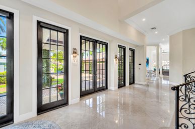 Sunlit luxury foyer in a tropical home with black-framed French doors and sidelights, glossy marble floors, wrought-iron staircase railing, wall sconces, and palm trees visible through the glass.