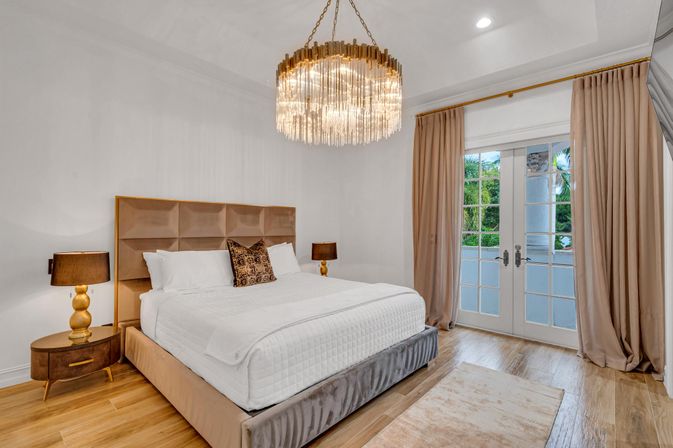 Bright luxury master bedroom with king bed and tufted headboard, crystal chandelier, wood floors, matching nightstands and French doors opening to a balcony with palm trees visible.