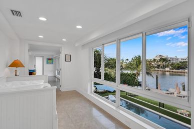 Sunlit white waterfront condo interior with large windows overlooking a pool, palm trees, canal, dock and blue sky.