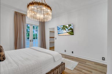 Bright modern luxury bedroom with crystal chandelier over a white-quilted king bed, French doors with beige curtains revealing palm trees, wall-mounted TV showing a beach scene, walk-in closet and wood floors