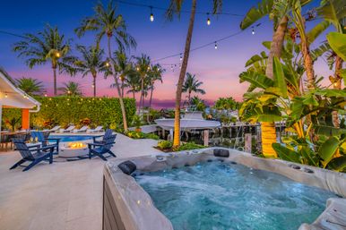 Twilight tropical waterfront patio with bubbling hot tub in foreground, pool and fire pit surrounded by blue Adirondack chairs, string lights, palm trees, and a boat dock on a calm canal.