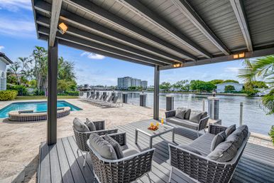 Covered waterfront patio with gray outdoor lounge seating, private pool and row of sun loungers overlooking a calm canal and palm trees.