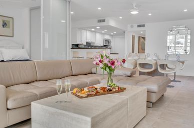 Sunlit modern open-plan living room and kitchen with beige leather sectional, marble coffee table holding a charcuterie board, two champagne flutes, and a vase of pink lilies near a white dining area.
