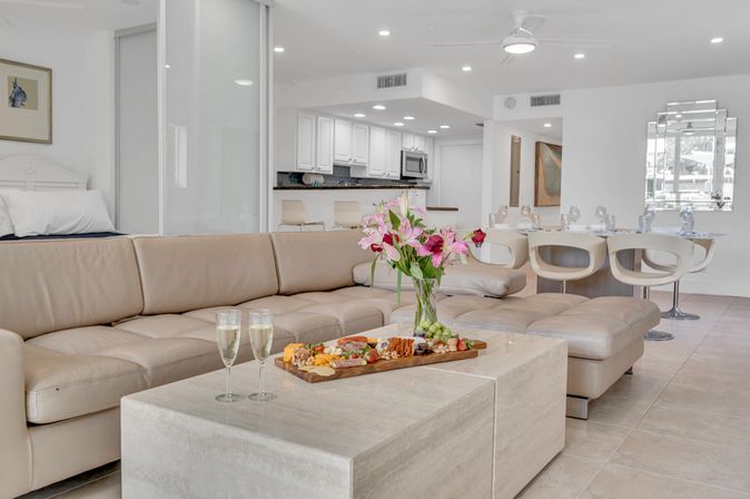 Sunlit modern open-plan living room and kitchen with beige leather sectional, marble coffee table holding a charcuterie board, two champagne flutes, and a vase of pink lilies near a white dining area.