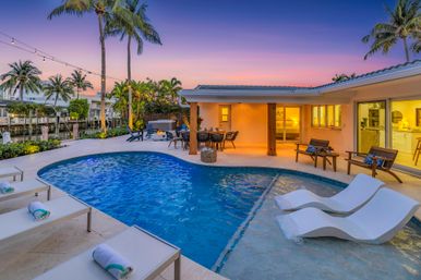 Sunset tropical waterfront backyard with curved pool, in-pool loungers, palm trees, string lights and canal-side outdoor dining.