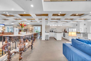 Bright open-concept clubhouse kitchen and lounge with white coffered ceiling, ornate carved wooden console table with floral centerpiece, long communal dining table, modern white kitchen islands and stainless appliances, and a blue sofa.