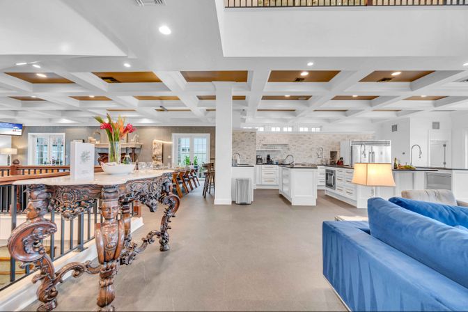 Bright open-concept clubhouse kitchen and lounge with white coffered ceiling, ornate carved wooden console table with floral centerpiece, long communal dining table, modern white kitchen islands and stainless appliances, and a blue sofa.