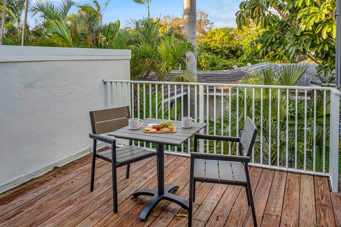 Cozy tropical balcony patio on a wooden deck with a small table, two chairs, coffee mugs and a fruit platter, white railing and lush palm trees beyond.