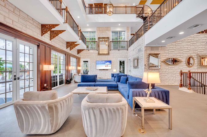 Bright coastal resort lounge with three-story atrium, stone walls, large navy-blue sectional, cream swivel chairs, balconies, and floor-to-ceiling glass doors opening to a waterfront patio.