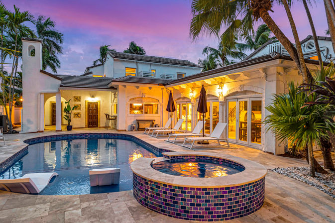 Twilight view of a luxury tropical backyard with a Mediterranean-style villa, illuminated patio, tiled swimming pool and circular hot tub, lounge chairs and umbrellas on a stone deck framed by palm trees and a pink‑purple sunset sky.