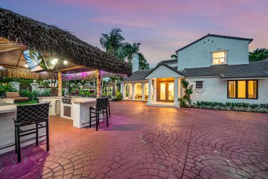 Luxury tropical backyard oasis at sunset with a thatched tiki bar and outdoor kitchen, red paver courtyard, palm trees and Mediterranean-style white house.