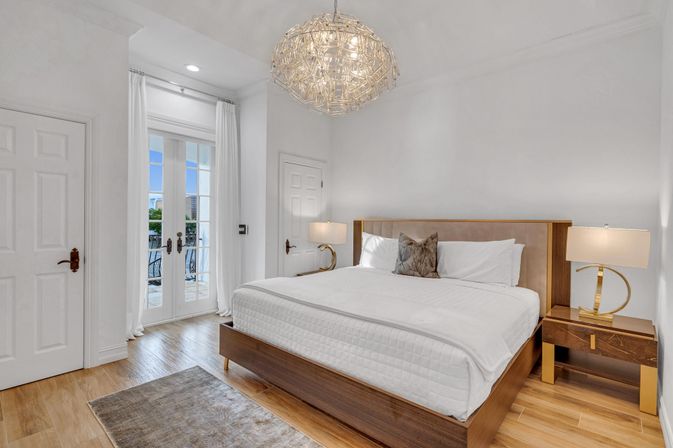 Bright modern bedroom with king-size bed in white linens, walnut platform frame, matching nightstands and lamps, sculptural chandelier, French doors to balcony and light wood floors.
