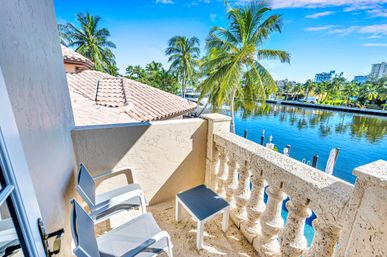Sunlit waterfront balcony with two white chairs and a small table overlooking a palm-lined canal, clay tile roofs and a distant coastal skyline under a bright blue sky