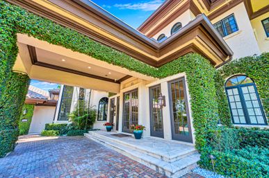Welcoming luxury Mediterranean-style home entrance with ivy-covered columns and overhang, arched windows, glass double doors, stone steps, potted plants, and a brick driveway under a bright blue sky.
