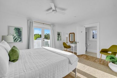 Sunlit modern bedroom with white quilted bed, green accent pillows, mid-century chairs and desk, and French doors opening to a balcony with palm trees — bright coastal retreat