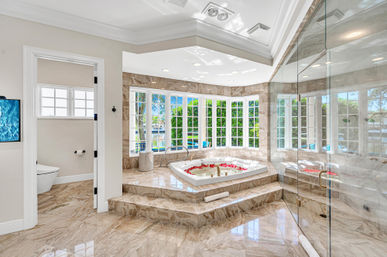 Bright luxury bathroom with polished marble floors and steps to a sunken soaking tub sprinkled with rose petals, surrounded by floor-to-ceiling bay windows with a leafy waterfront view, plus a glass walk-in shower and separate toilet room.