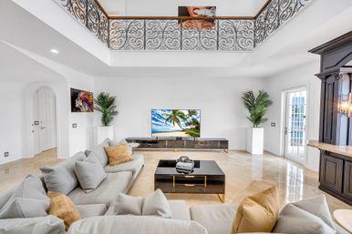 Bright luxury living room with double-height ceiling and ornate wrought-iron balcony, neutral sectional sofa with tan pillows, black coffee table, TV showing a beach, marble floors and potted palms.