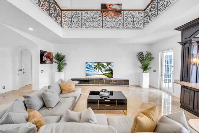 Bright luxury living room with double-height ceiling and ornate wrought-iron balcony, neutral sectional sofa with tan pillows, black coffee table, TV showing a beach, marble floors and potted palms.
