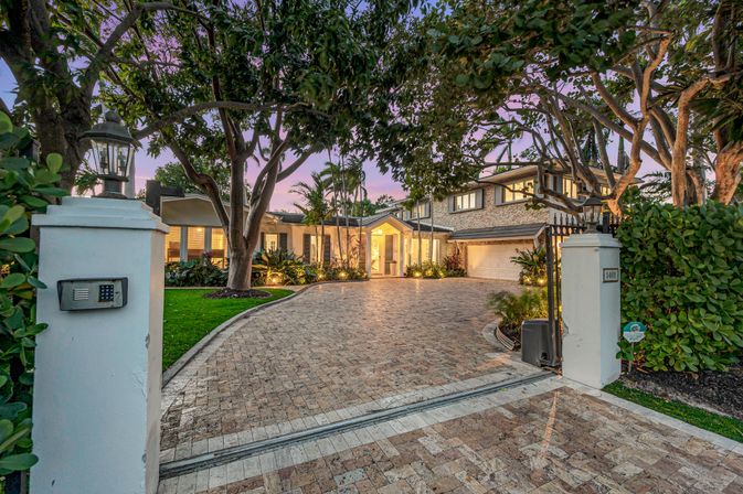 Gated paver driveway framed by mature trees and palms leading to a warmly lit two-story luxury home at dusk, with manicured landscaping and lantern-topped pillars.