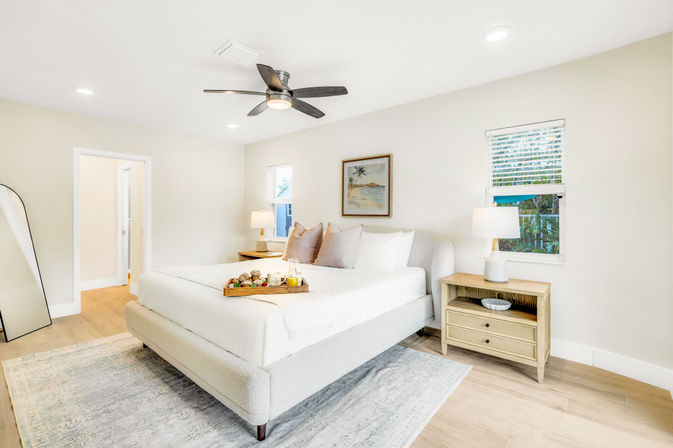 Sunlit coastal-style bedroom with king-size upholstered bed, neutral linens, wooden nightstands and lamps, ceiling fan, breakfast tray on bed, light wood floors and area rug.